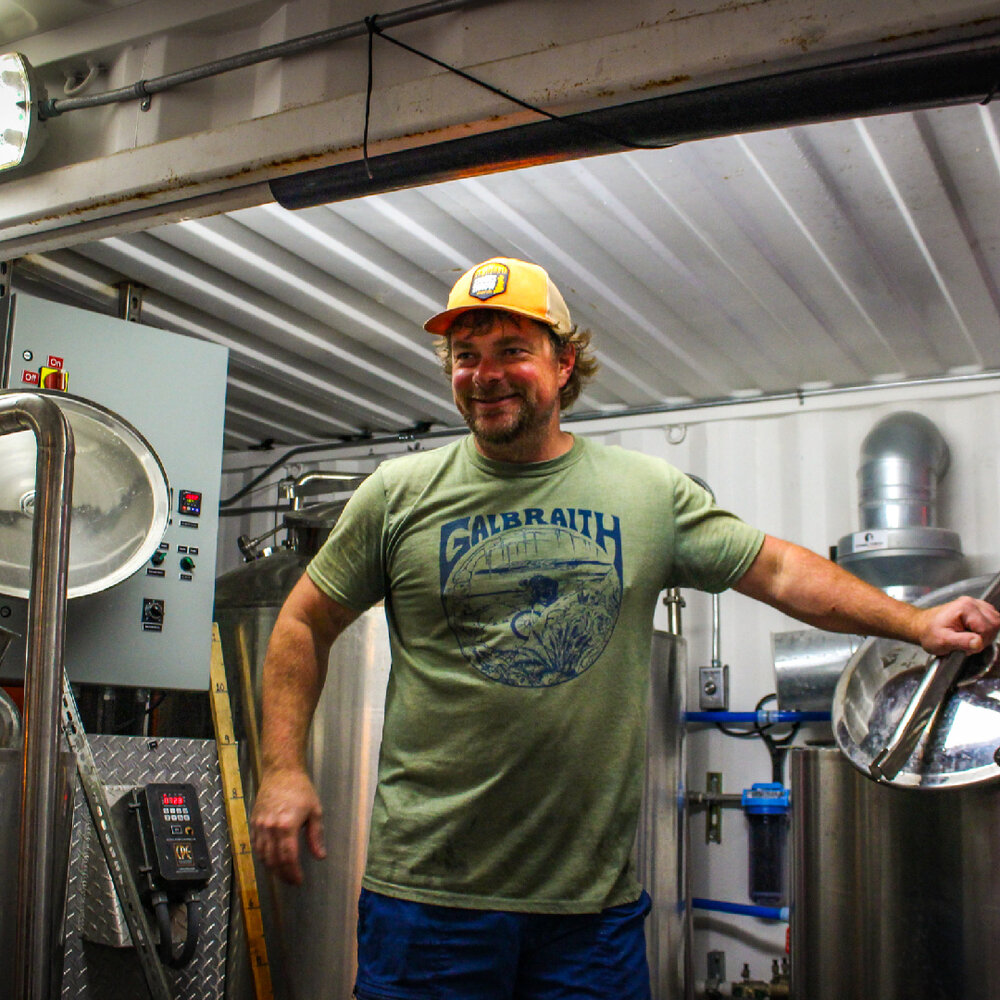 A man in the working part of a brewery smiles near the stainless casks.